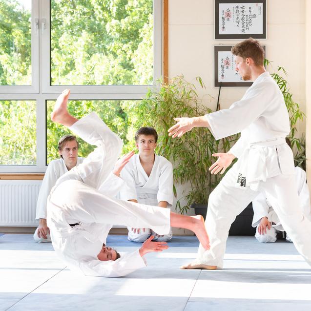 Zwei Judokas in weißen Gi-Shirts, einer führt eine Wurftechnik aus, während andere Zuschauer aufmerksam beobachten.