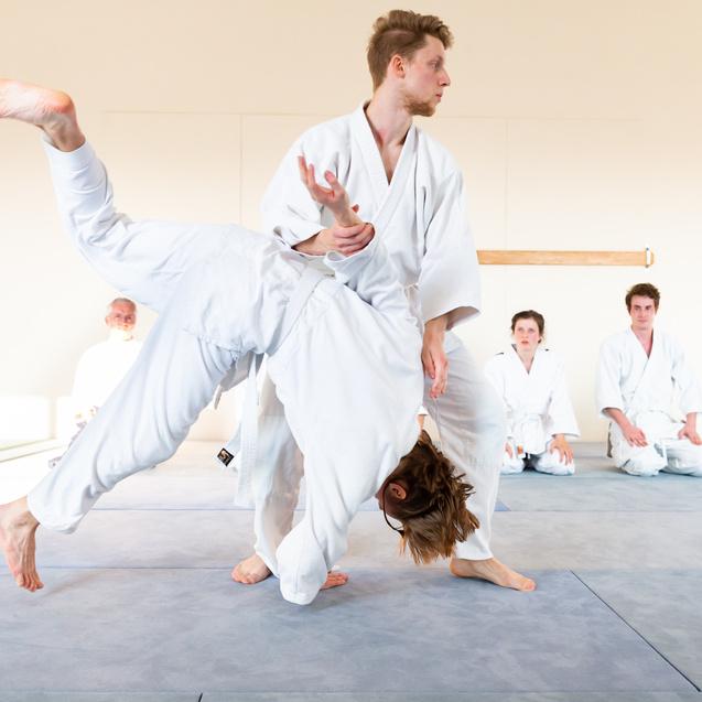 Zwei Kampfsportler in weißen Anzügen führen eine Wurftechnik auf einer Matte aus, während andere zuschauen.