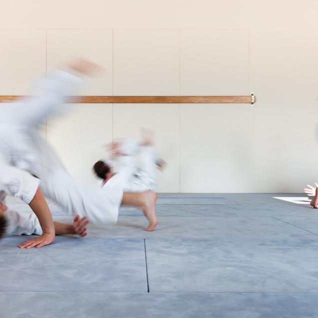 Drei Kinder in weißen Anzügen führen akrobatische Bewegungen auf einer blauen Matte in einem hellen Raum aus.