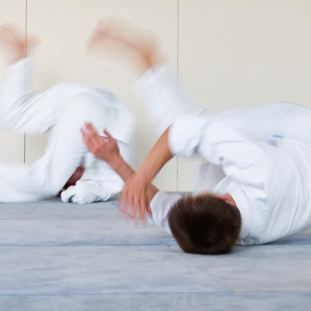 Zwei Judoka in weißen Gi rollen auf einer Matte, Bewegungen sind verschwommen und dynamisch.