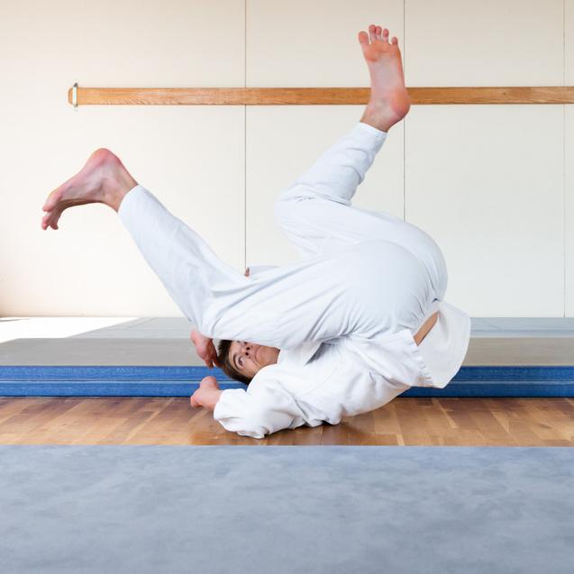 Trainierende in weißen Judokis üben akrobatische Bewegungen auf einer Matte, mit einer weiteren Person im Hintergrund.