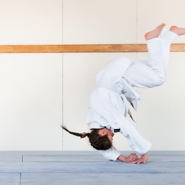 Mädchen in Karate-Anzug führt einen Kopfstand auf einer blauen Matte in einem Trainingsraum aus.