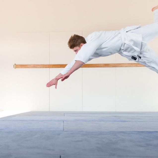 Springender Kampfsportler in weißem Gi auf einer Matte, mit gestreckten Armen in der Luft.