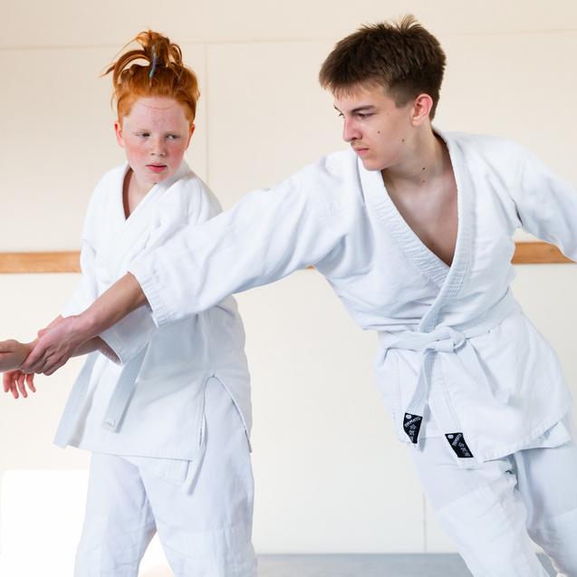 Zwei Jugendliche in weißen Judoanzügen üben einen Griff in einem hellen Trainingsraum.