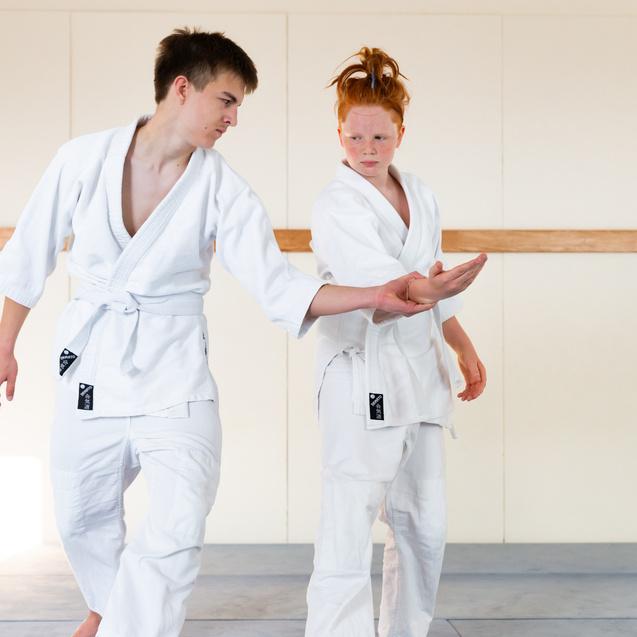 Zwei Jugendliche in weißen Judogis üben einen Bewegungsablauf auf einer Judomatte in einem hellen Raum.