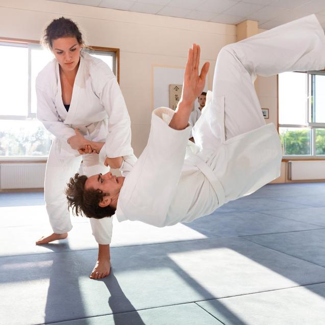 Junge Judoka in Weiß führt einen Wurf an einem Partner durch, der sich in der Luft befindet. Judohalle im Hintergrund.