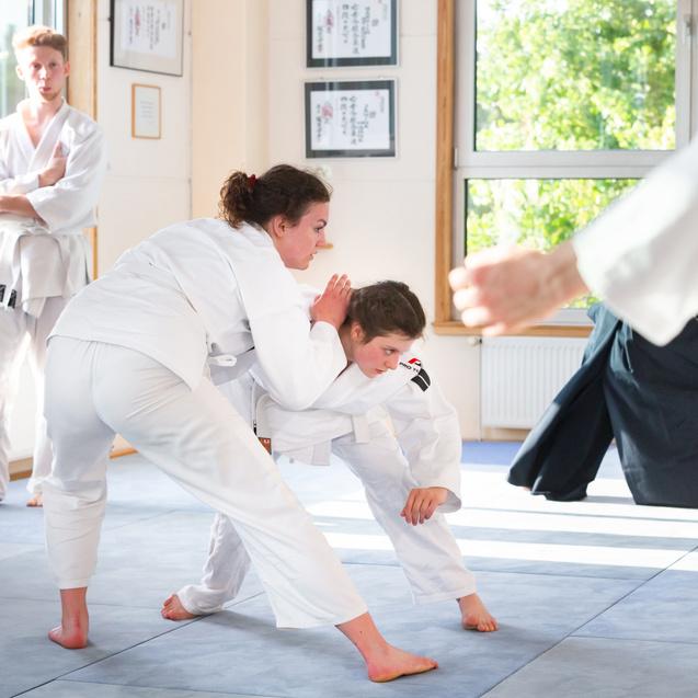 Zwei Personen im weißen Judo-Gi üben Grappling-Techniken auf einer blauen Judo-Matte in einem hellen Raum.