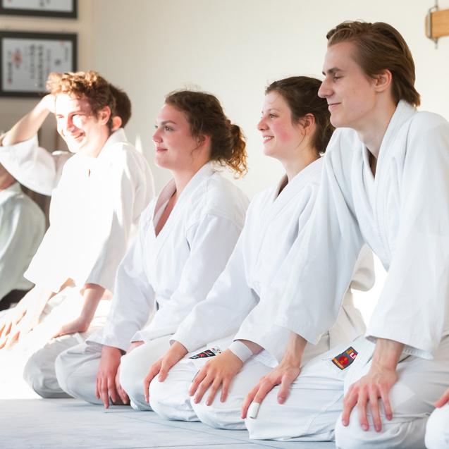 Sechs Personen in weißen Judoge robes sitzen lächelnd auf dem Boden in einem hellen Raum mit Holzständern.