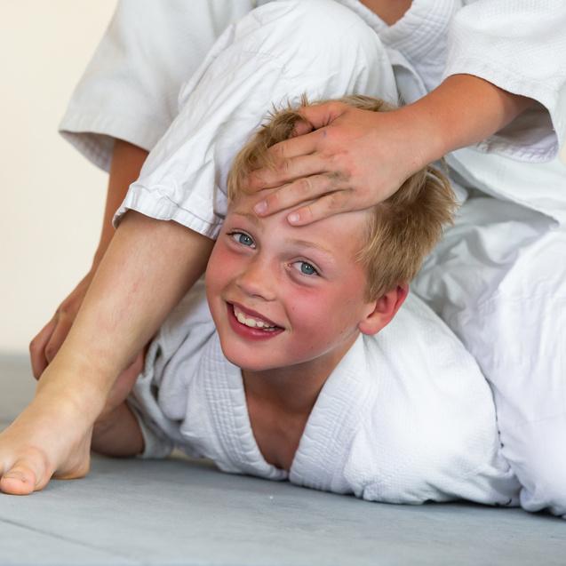 Junge in Judoanzug, lacht während eines Trainings, während ein Partner ihn von oben hält.