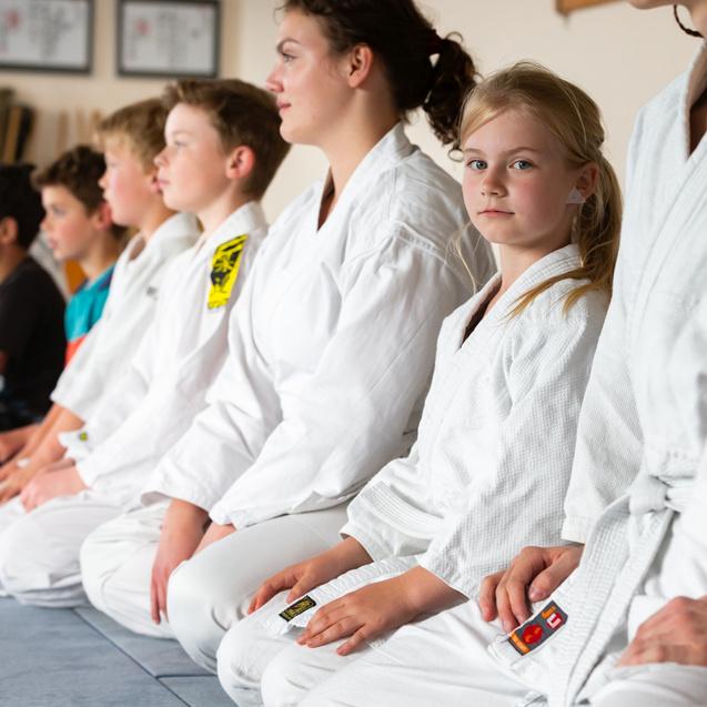 Gruppe von Kindern und Jugendlichen in weißen Trainingsanzügen, sitzend auf einer Matte im Dojo.