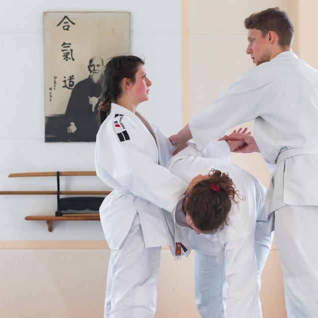 Drei Personen in weißen Judogis üben Techniken in einem Trainingsraum mit einer Wand als Hintergrund.