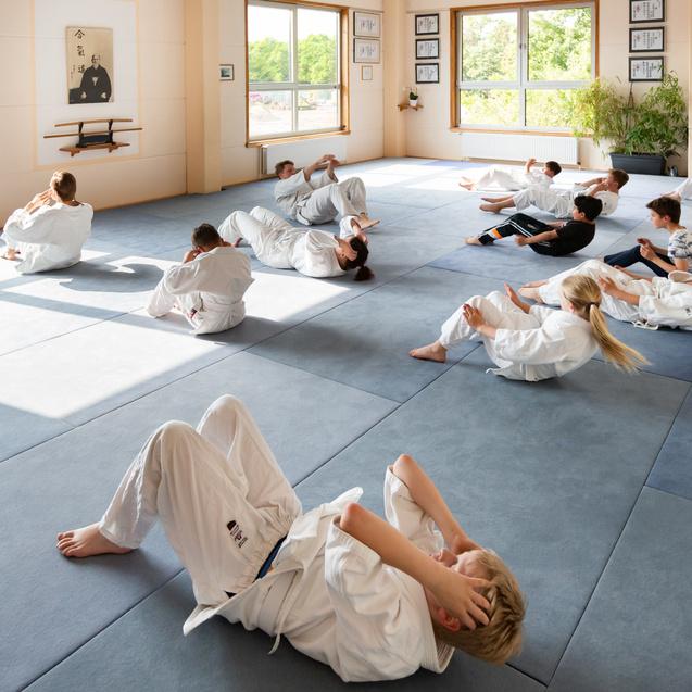 Gruppe von Kindern in weißen Judoanzügen, die auf blauen Matten Bauchübungen im Sonnenlicht durchführen.