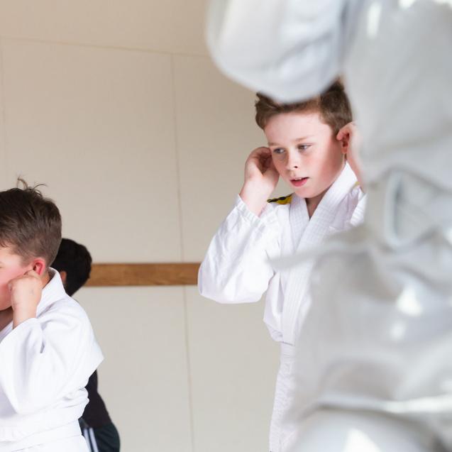 Zwei Kinder in weißen Judoanzügen üben in einem Dojo, während ein weiterer Teilnehmer im Hintergrund sichtbar ist.