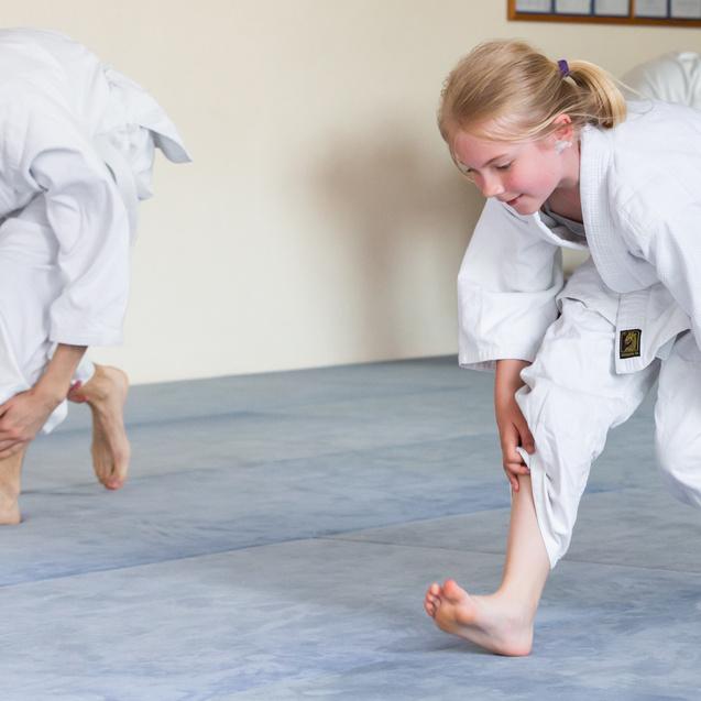 Zwei Kinder in weißen Judo-Anzügen üben Bewegungen auf einer Judo-Matte in einem Übungsraum.