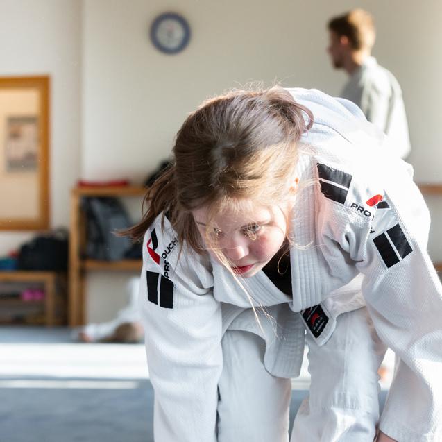 Junge in Judokleidung beugt sich auf einer Matte, während ein weiterer junger Mensch im Hintergrund steht.