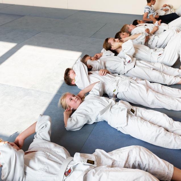Gruppe von Kindern in weißen Judo-Anzügen liegt auf blauen Matten in einem hellen Raum.