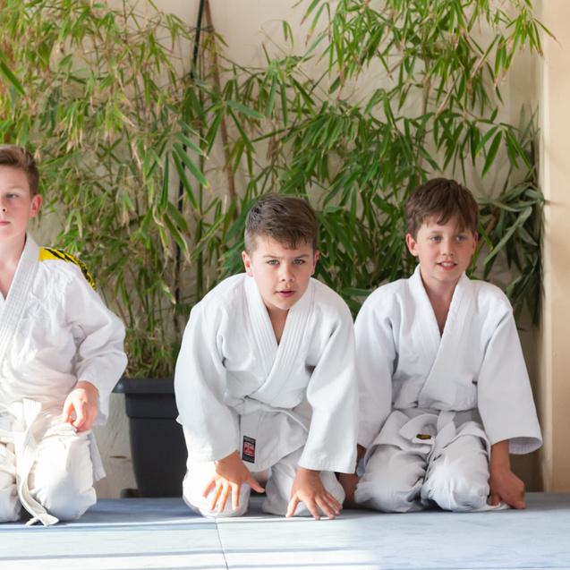 Vier Kinder in weißen Judoanzügen sitzen auf einer Matte, umgeben von Pflanzen im Hintergrund.