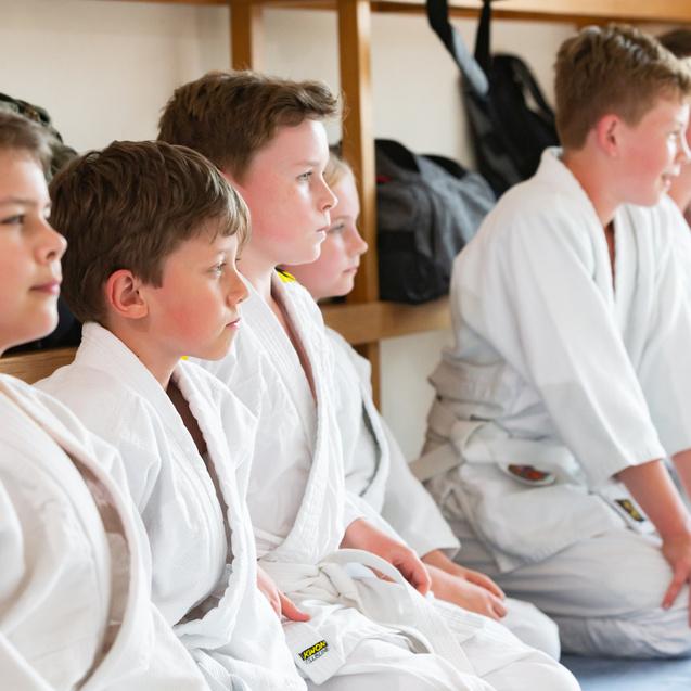 Sechs Kinder in weißen Judokimonos sitzen in einer Reihe auf dem Boden in einem Trainingsraum.