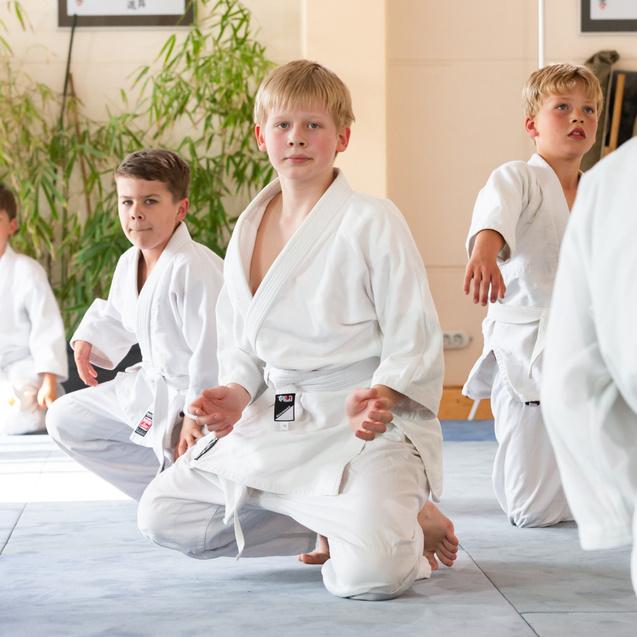 Gruppe von vier Kindern in weißen Judoanzügen, die auf einer Matte in einem Trainingsraum üben.
