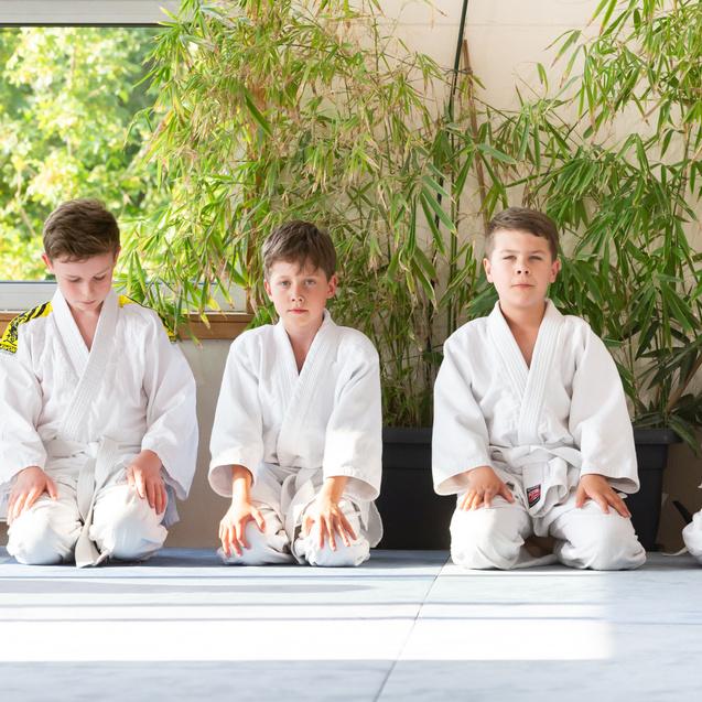 Fünf Kinder in weißen Judo-Anzügen sitzen auf einer Matte vor Fenstern mit Pflanzen im Hintergrund.