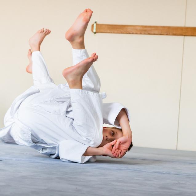 Ein Kind in weißem Judogi rollt auf einer blauen Matte, während ein anderes Kind im Hintergrund beobachtet.
