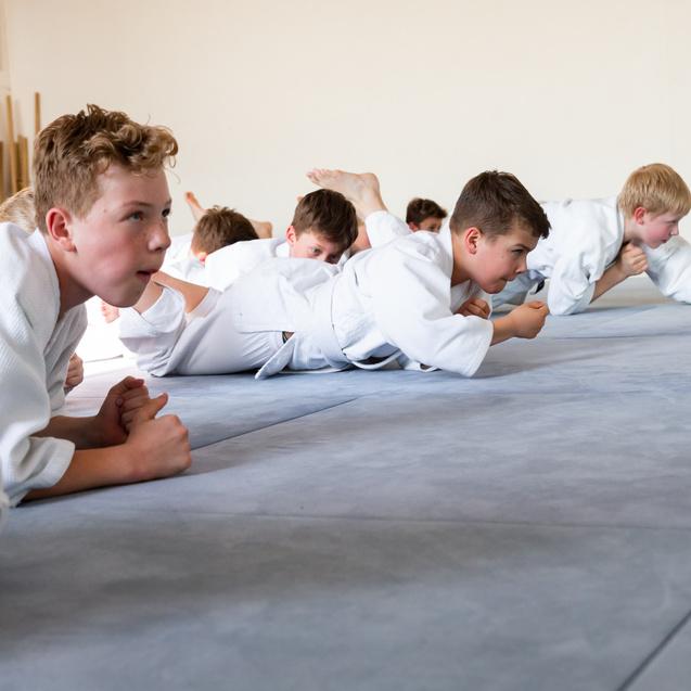 Gruppe von Kindern in weißen Judoanzügen übt liegend auf einer blauen Matte in einem Trainingsraum.