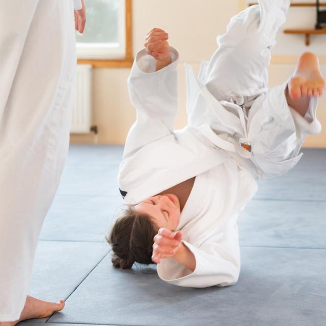 Junge Kämpferin in weißem Gi führt einen Wurf auf einer gepolsterten Matte aus.