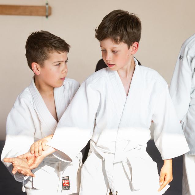 Zwei Jungen in weißen Judogis üben einen Griff und stehen auf einer Judomatte in einem Trainingsraum.