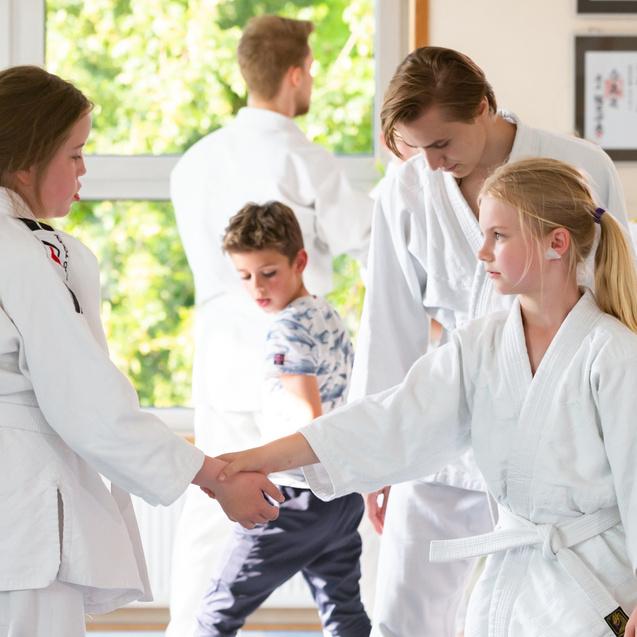 Zwei Kinder im Judogi schütteln sich die Hände in einem Trainingsraum, während weitere Kinder im Hintergrund aktiv sind.