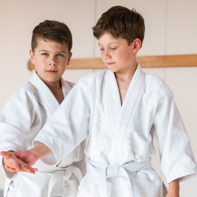 Zwei Jungen in weißen Judo-Anzügen üben Techniken in einem Trainingsraum mit Holzblende im Hintergrund.