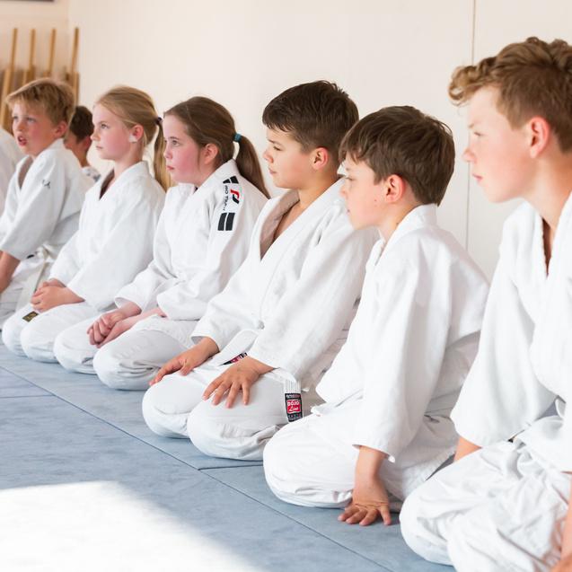 Gruppe von Kindern in weißen Judogi, die auf einer Matte sitzen und konzentriert nach vorne blicken.