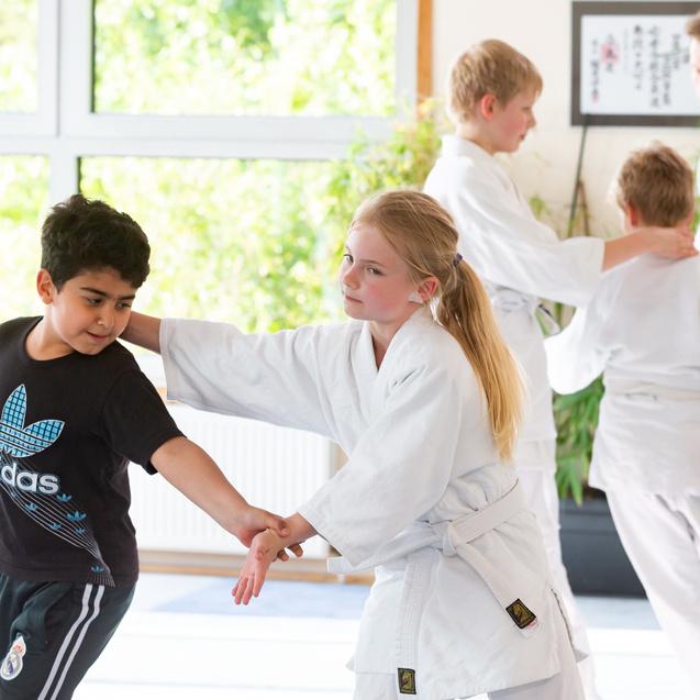 Zwei Kinder im Judoanzug üben Partnertechniken in einem Raum mit großen Fenstern und Pflanzen im Hintergrund.