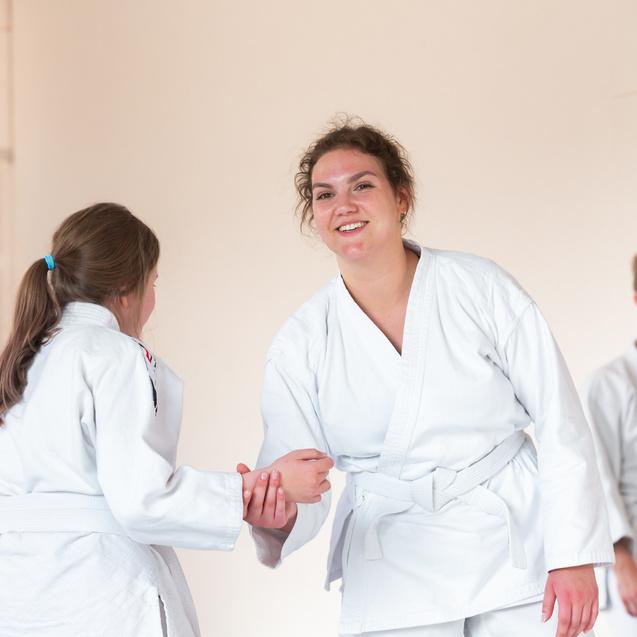 Zwei Trainingsteilnehmerinnen im BJJ-Kampfanzug begrüßen sich freundlich in einem Dojo, weitere Schüler im Hintergrund.