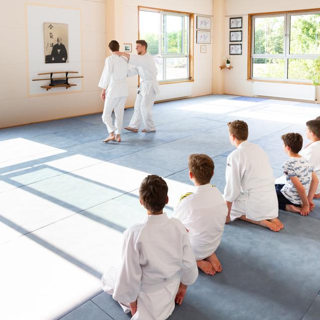 Zwei Erwachsene in Kampfsportanzügen demonstrieren Techniken vor einer Gruppe sitzender Kinder im Dojo.