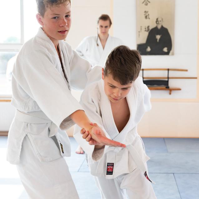 Zwei Kinder im Judoanzug üben zusammen eine Technik in einem Trainingsraum mit matten und Wanddekoration.
