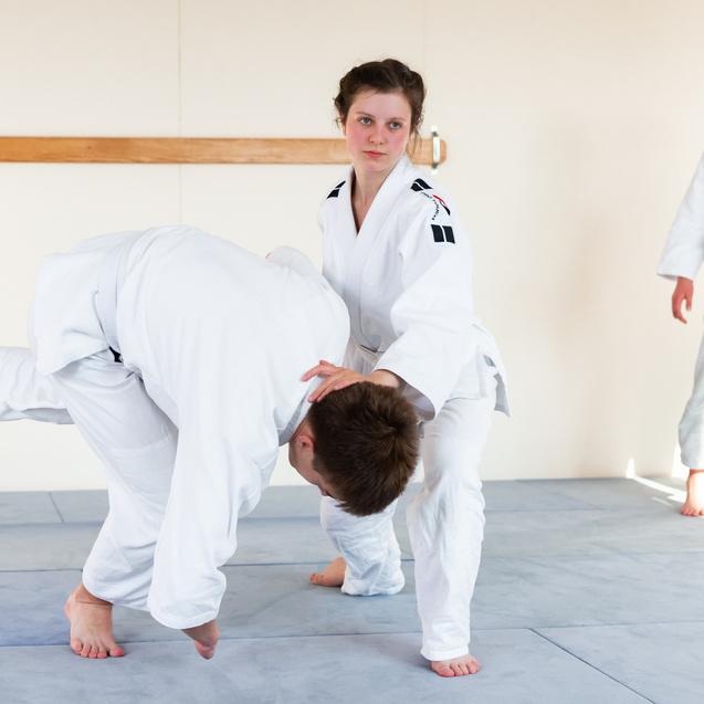 Zwei Judokas trainieren einen Wurf in einem hellen Trainingsraum, während eine dritte Person zusieht.