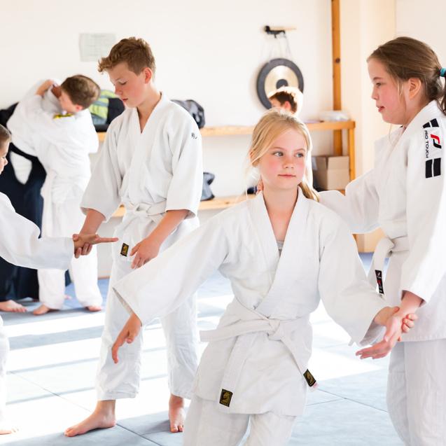 Vier Kinder in weißen Judogis üben Partnerübungen in einem hellen Trainingsraum mit Matten.