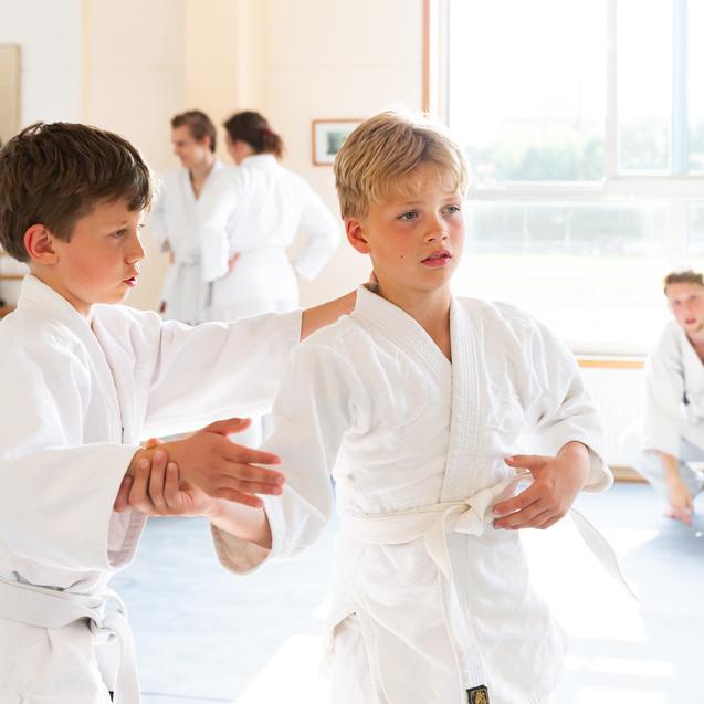 Zwei Jungen in weißen Judoanzügen üben im Dojo, während andere Kinder im Hintergrund aktiv sind.