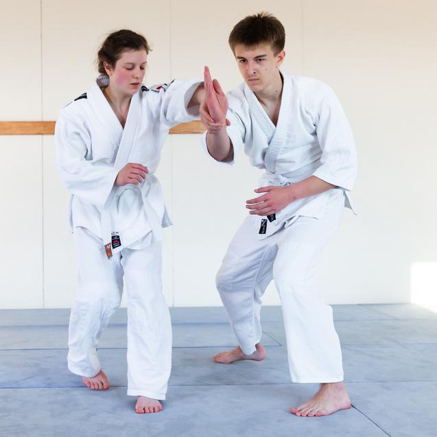 Zwei Jugendliche in weißen Judoanzügen üben eine Technik auf einer Judomatte in einem Trainingsraum.