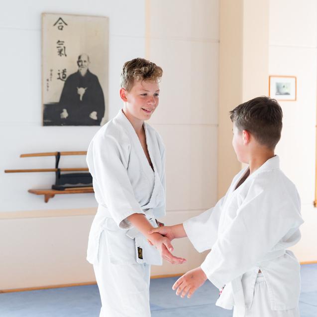 Zwei Jungen in weißen Judogi üben eine Technik im Dojo vor einem traditionellen Wandbild.
