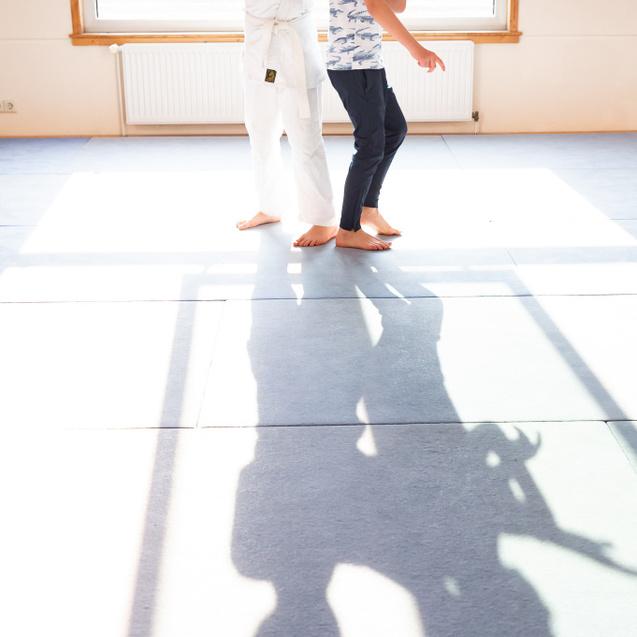 Zwei Jungen im Training: einer im weißen Gi zeigt einer anderen Person eine Technik, während sie in einem lichtdurchfluteten Raum stehen.