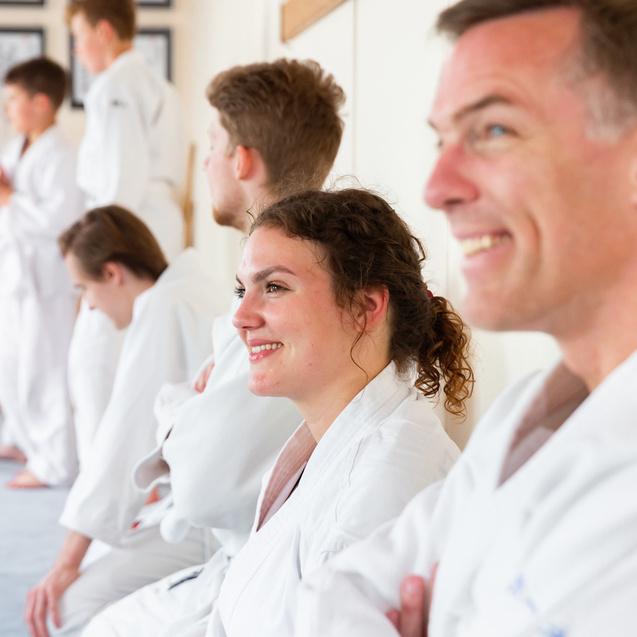 Gruppe junger Kampfsportler in Weiß, lächelnd und wartend, während andere im Hintergrund trainieren.