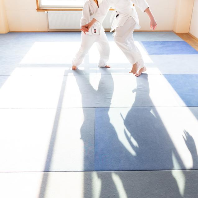 Zwei Jungen in weißen Karateanzügen üben Bewegungen auf einer blauen Matte im hellen Trainingsraum.