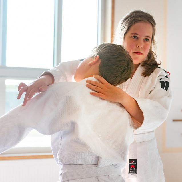 Zwei Kinder in Judoanzügen üben eine Technik in einem hellen Raum mit Fenster und Judo-Dekoration.