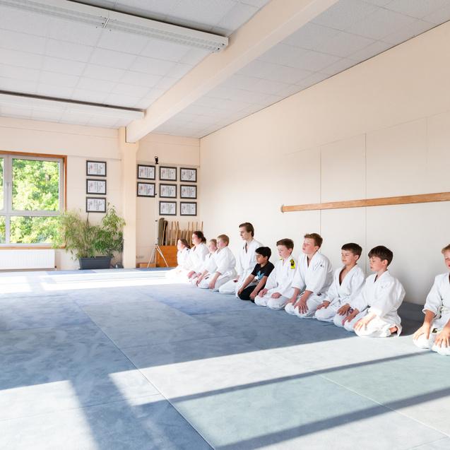 Junge Martial-Arts-Schüler in weißen Anzügen sitzen auf einer Matte, während ein Trainer vor ihnen meditierend kniet.