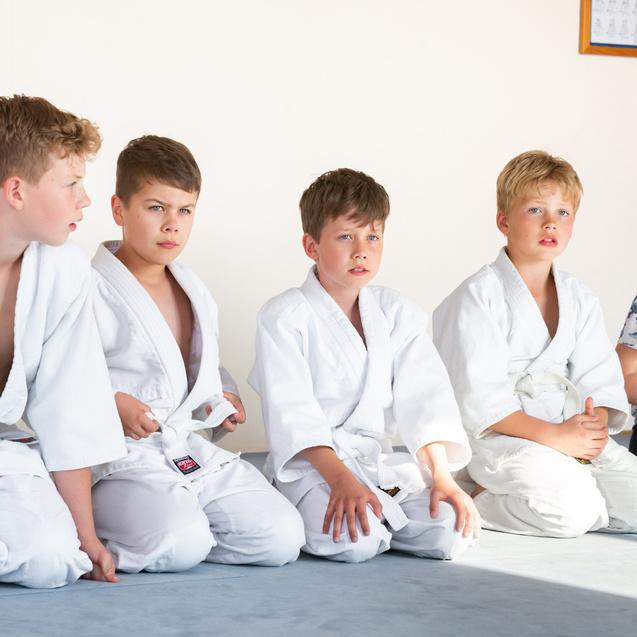 Fünf Kinder in weißen Judoanzügen sitzen auf dem Boden und schauen aufmerksam nach vorne.