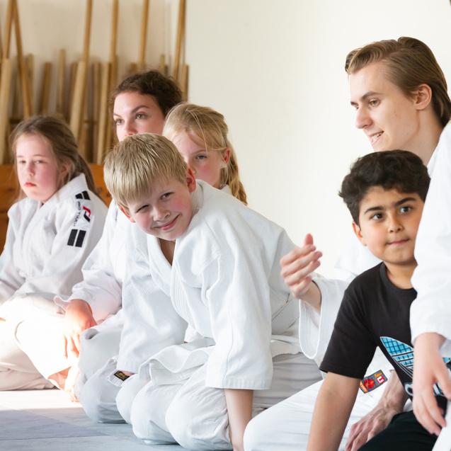 Gruppe von Kindern sitzt auf der Matte, einige in Judoanzügen, andere in Alltagskleidung, während sie einer Person zuhören.