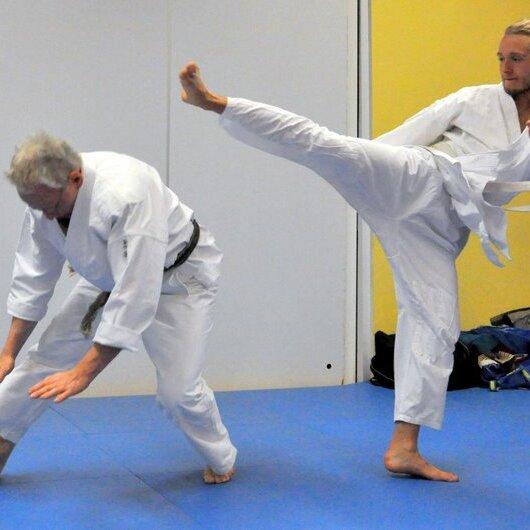 Zwei Judoka in weißen Anzügen kämpfen auf einer blauen Matte; einer führt einen hohen Kick aus, der andere weicht aus.