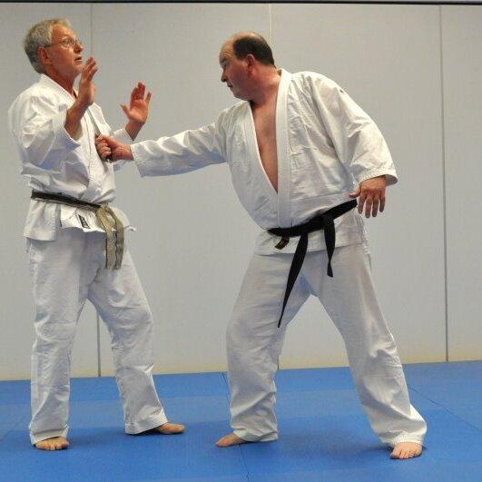 Zwei Männer in weißen Judoanzügen üben eine Technik auf einer blauen Judomatte.