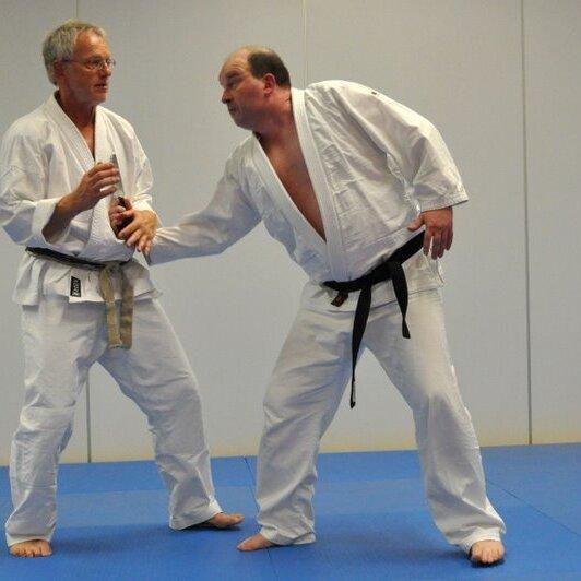 Zwei Judoka in weißem Gi stehen auf einer blauen Matte und üben eine Technik. Ein Partner hat einen schwarzen Gürtel.